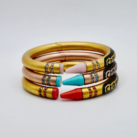 Three stacked bangle bracelets designed to look like crayons, featuring gold-colored bodies with rhinestone accents and colored tips in red, turquoise, and light pink, on a light gray background.