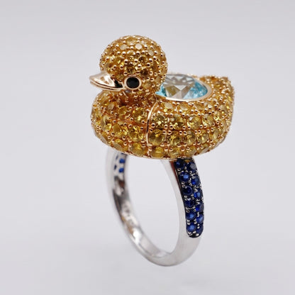 A sculptural ring shaped like a duck, featuring a textured gold duck body and head, a light blue oval gemstone on its back, a small black eye, and a silver band accented with dark blue pavé stones.